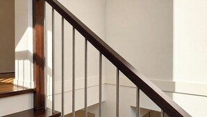 Una escalera de madera con barandilla de vidrio, que conduce a un rellano con pared blanca y piso de madera.