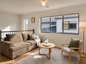 A living room with a sofa, a chair, a coffee table, and a window with a view of a house. - 4BR Point Cook Home | Sleeps 8 | Free Parking (Point Cook)