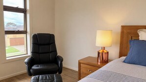 Un dormitorio con una cama, una mesita de noche con lámpara, un sillón de cuero negro y un ventanal con vistas a un edificio y árboles.