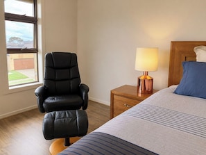 A bedroom with a bed, a nightstand with a lamp, a black leather chair, and a window with a view of a building and trees. - 4BR Point Cook Home | Sleeps 8 | Free Parking (Point Cook)