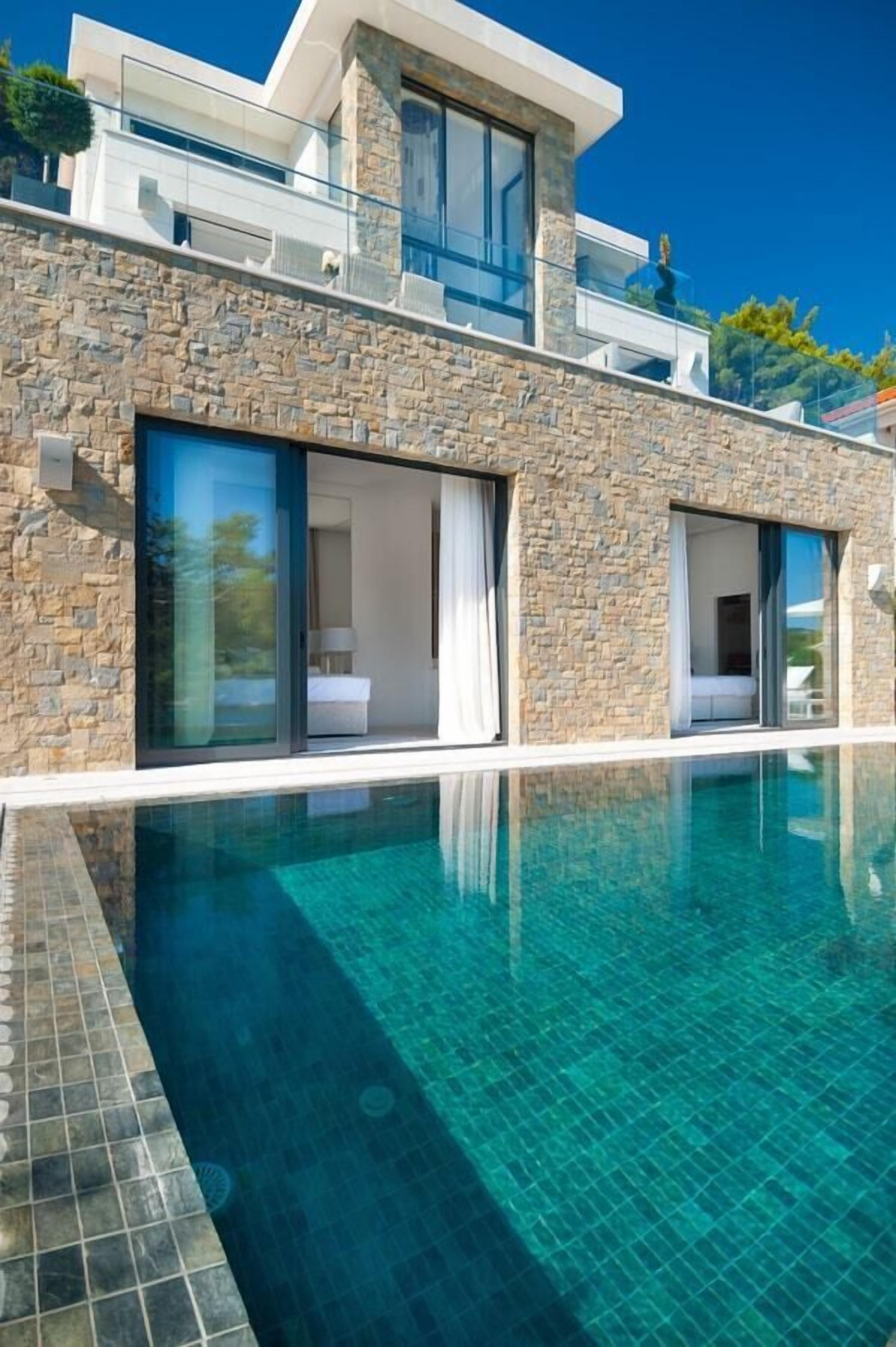 Un edificio moderno con fachada de piedra, grandes puertas correderas de cristal y una piscina azul cristalina.