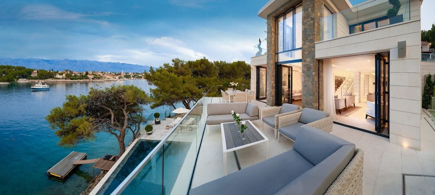 Une maison moderne dotée d’une piscine à débordement en verre donnant sur un lac, d’un coin repas et offrant une vue sur les montagnes.