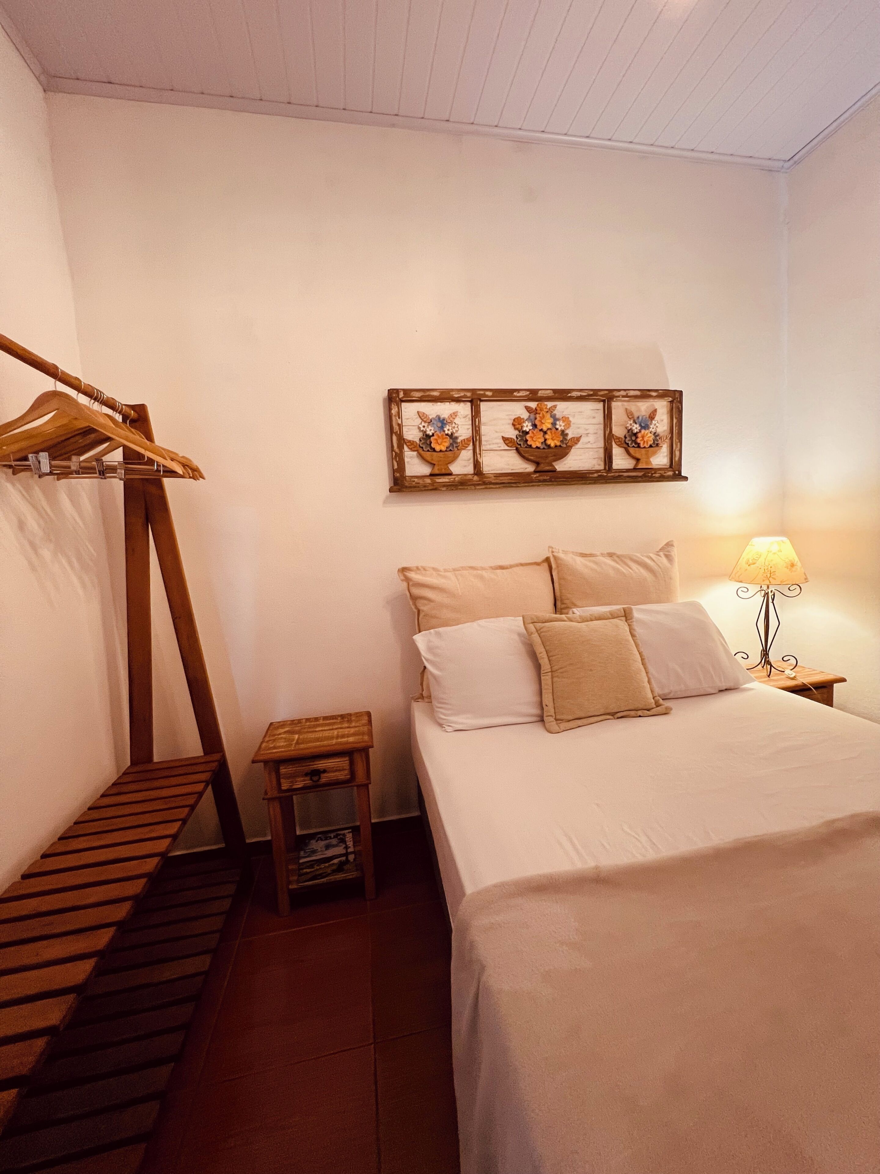 Un dormitorio con cama, un perchero de madera, una mesita de noche pequeña de madera y un cuadro colgado en la pared.