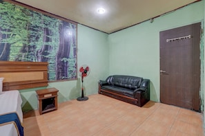 A room with a bed, a wooden dresser, a black leather sofa, a standing fan, a door, and a wall with a forest mural.