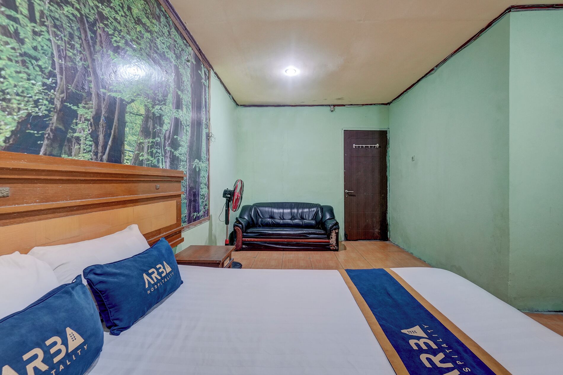 A hotel room with a bed, a wooden headboard, a blue pillow, a black leather sofa, a door, and a wall with a forest mural.