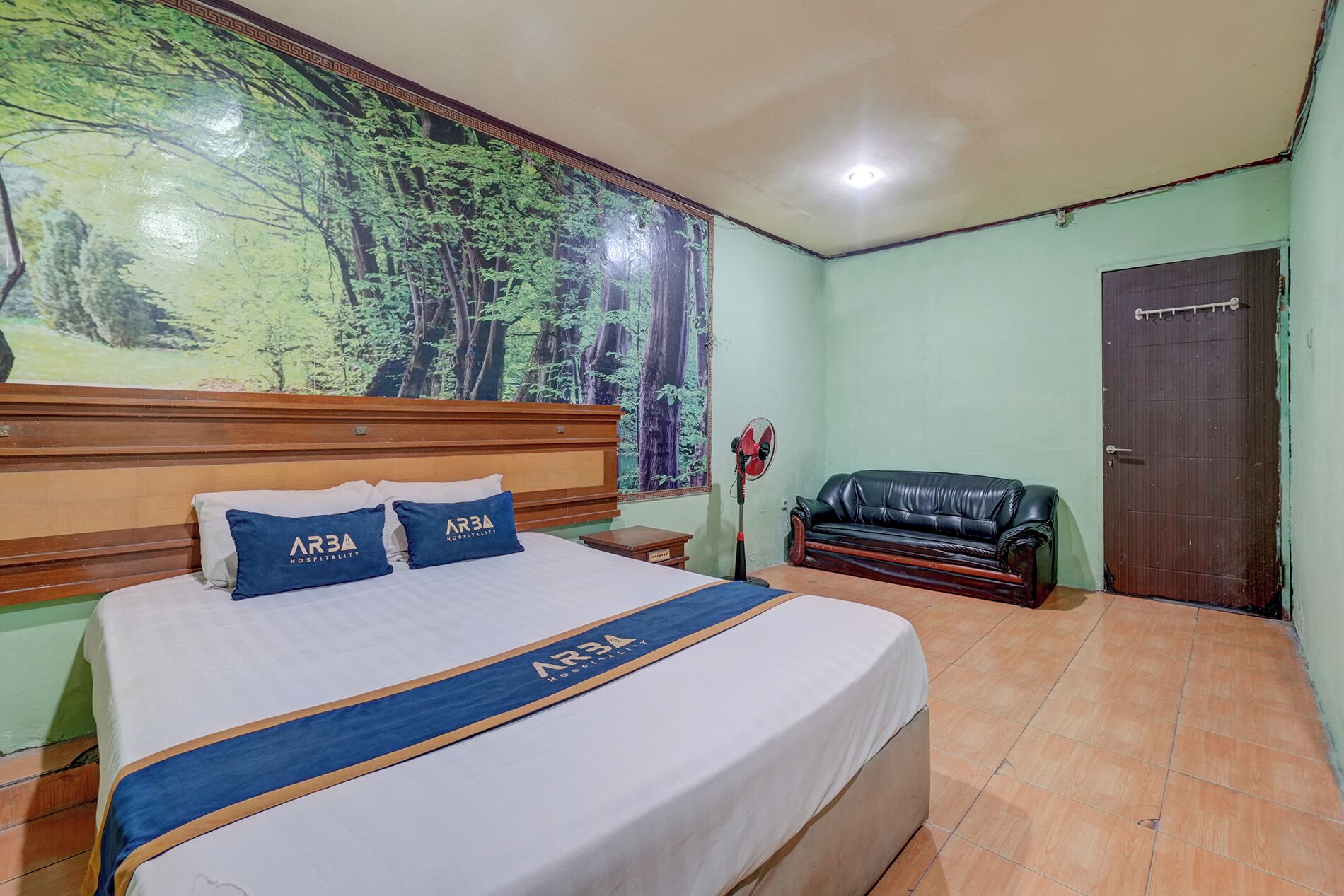 Quarto de hotel com uma cama grande, cabeceira, painel na parede, sofá de couro preto, mesa de cabeceira de madeira e uma porta.