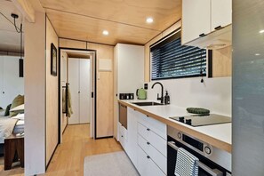 A compact kitchen with a built-in oven, a sink, and a window with blinds. - Kakariki Tiny House - Bush Sauna, Okere Falls (Rotorua)