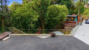 A rooftop with a glass balustrade, a small garden area with potted plants, and a view of a forested area.