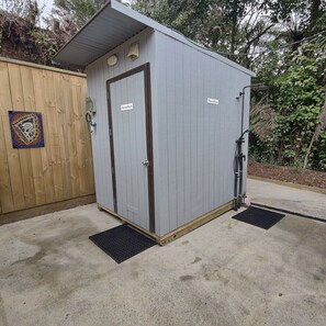 Un cobertizo pequeño de un piso con una puerta y una ventana, ubicado al lado de una cerca de madera y un camino de concreto.