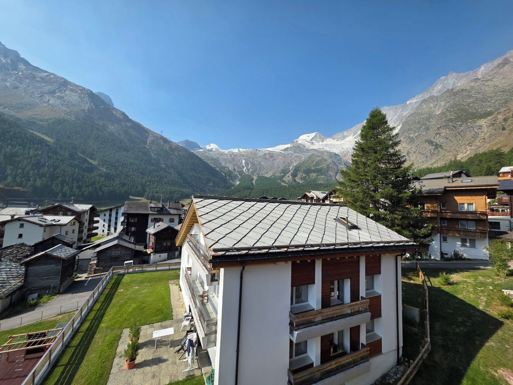 A mountain village with traditional wooden houses, a modern building with a sloped roof, and a clear view of snow-capped mountains.
