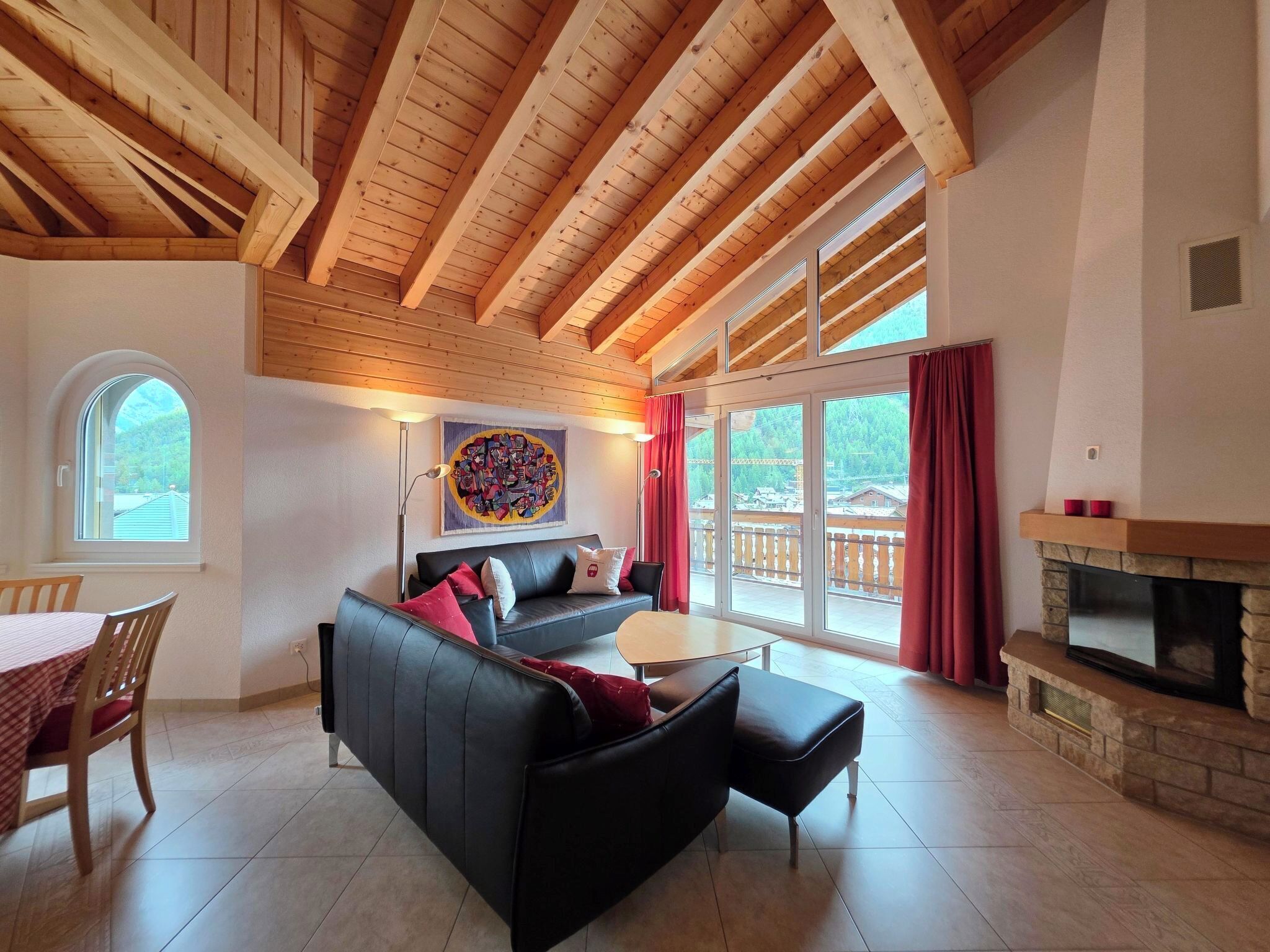 A spacious living area with a black sofa, a dining table with chairs, a fireplace, and large windows offering a view of the outdoors.