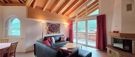 A spacious living area with a black sofa, a dining table with chairs, a fireplace, and large windows offering a view of the outdoors.