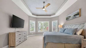 A bedroom with a bed, a television, a ceiling fan, and windows with blinds.