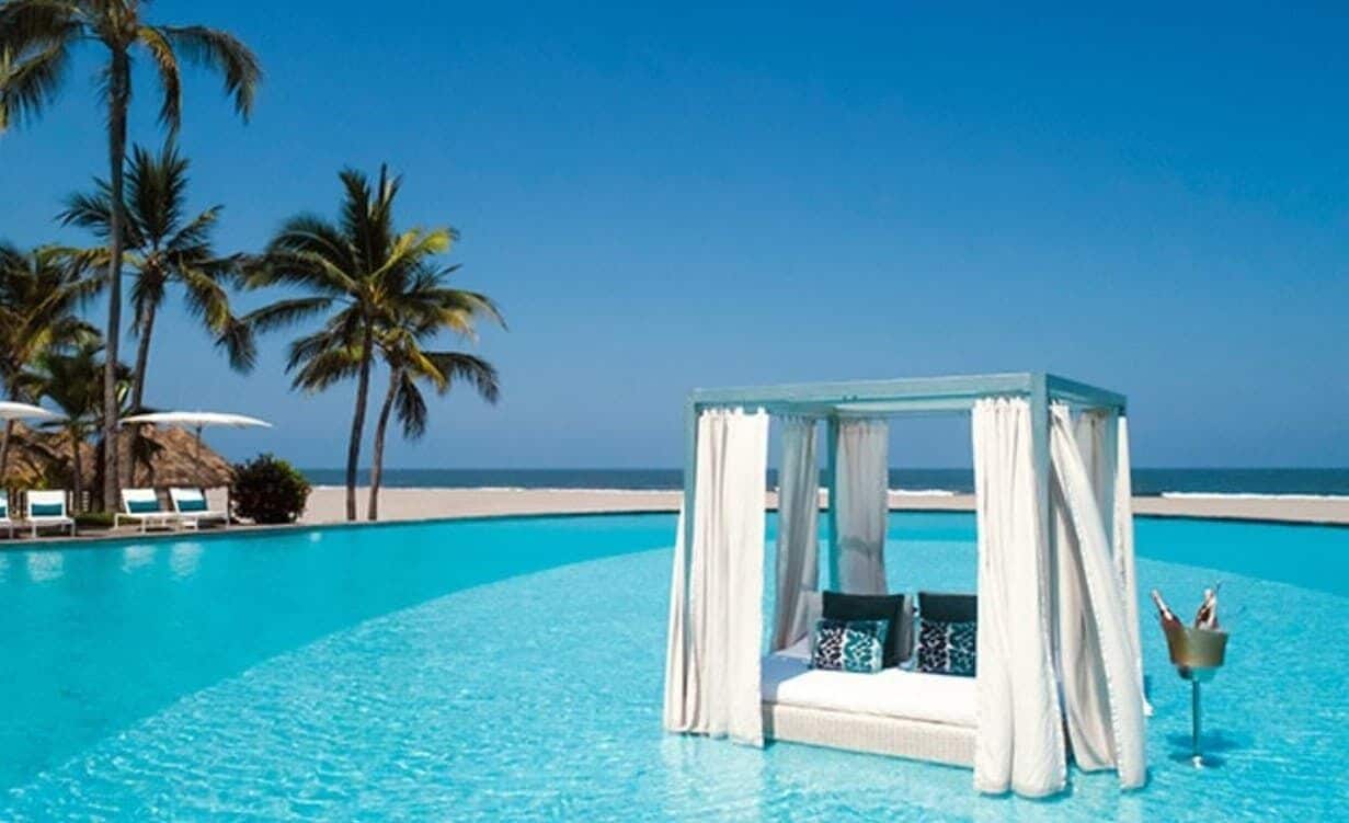 Ein klares, blaues Schwimmbecken mit einem weißen Gazebo unter einer Markise, Palmen und ein Strand im Hintergrund.