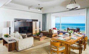 Ein geräumiges Wohnzimmer mit einem großen Flachbildfernseher, bequemen Sitzgelegenheiten, einem Essbereich mit Tisch für vier Personen und einem Balkon mit Meerblick.