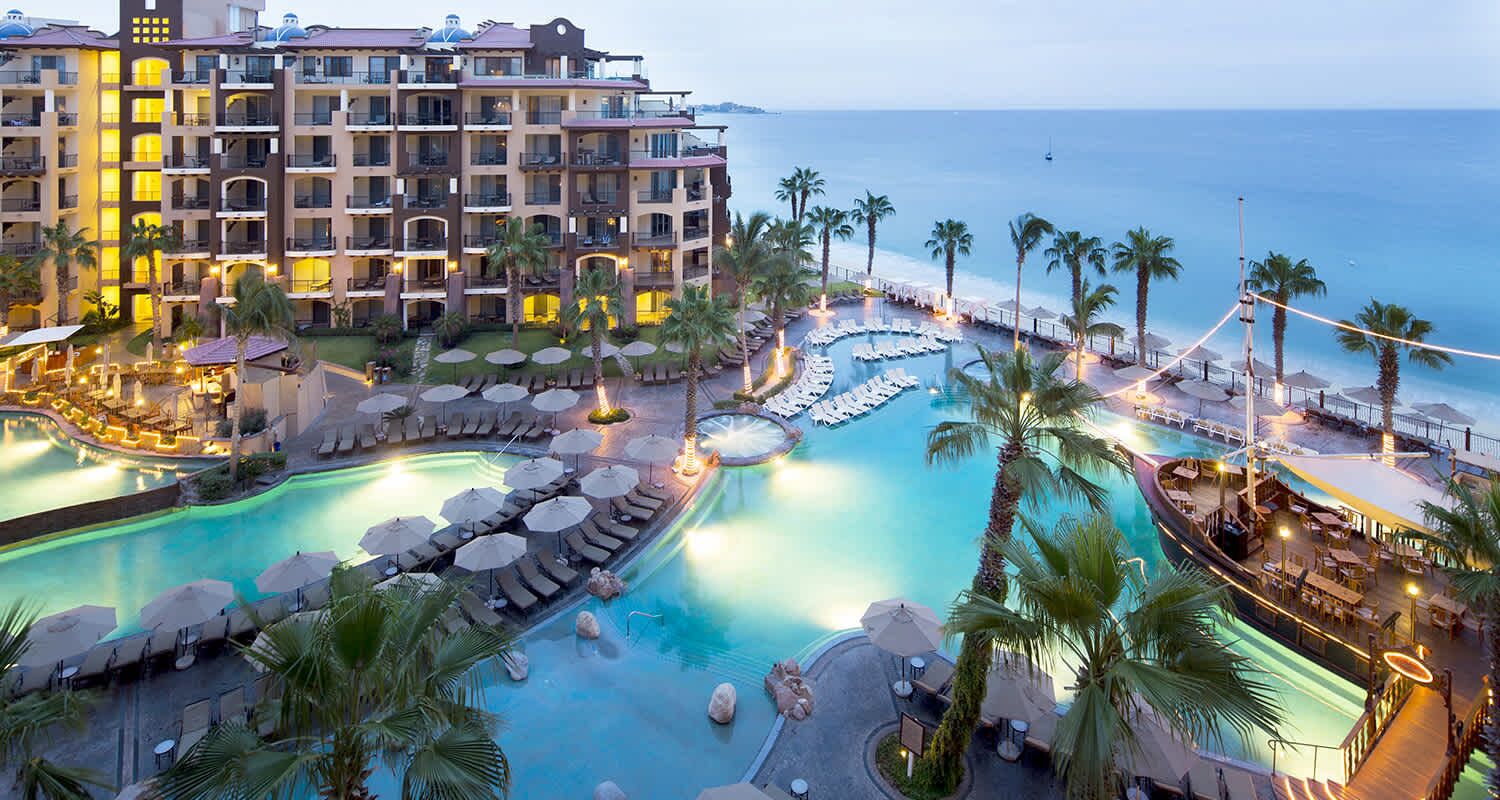 A hotel with multiple swimming pools, palm trees, and a view of the ocean.
