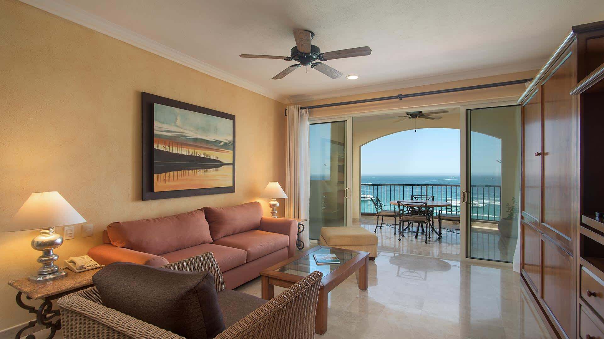 A living room with a sofa, a coffee table, and a painting on the wall. There is a balcony with a view of the ocean.