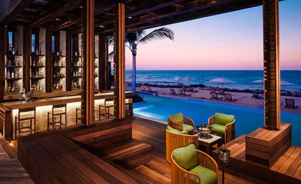 Eine Bar mit Holzboden und Sitzgelegenheiten am Pool mit Blick auf einen Strand bei Sonnenuntergang.