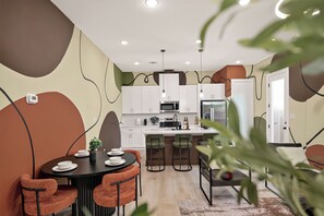 A modern kitchen with white cabinets, a central island, and a dining area with orange chairs. - Stunning Designer Home Minutes to NRG, Medical Center & Downtown Sleeps 9 (Houston)