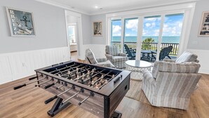 A room with a foosball table, a seating area with chairs, and a view of the ocean through large windows. - Sea Y'all Soon - Oceanfront in Garden City Beach (Garden City Beach)