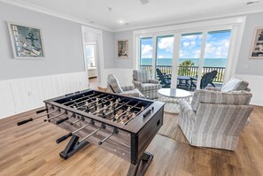 Ein Zimmer mit einem Kicker, einer Sitzecke mit Stühlen und Meerblick durch große Fenster.