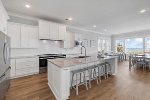 Een moderne keuken met witte kasten, een centraal eiland met barkrukken en een uitzicht op de omgeving door grote ramen.