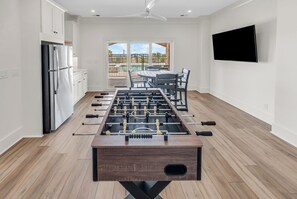 A modern kitchen with a foosball table, a dining area with a pool view, and a flat-screen TV mounted on the wall. - Coastal Point of View 10 Bedroom - Oceanfront in Garden City Beach (Garden City Beach)