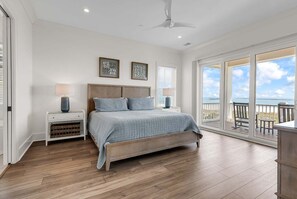A bedroom with a large bed, two bedside tables, a ceiling fan, and a view of the ocean through large windows. - Coastal Point of View 10 Bedroom - Oceanfront in Garden City Beach (Garden City Beach)