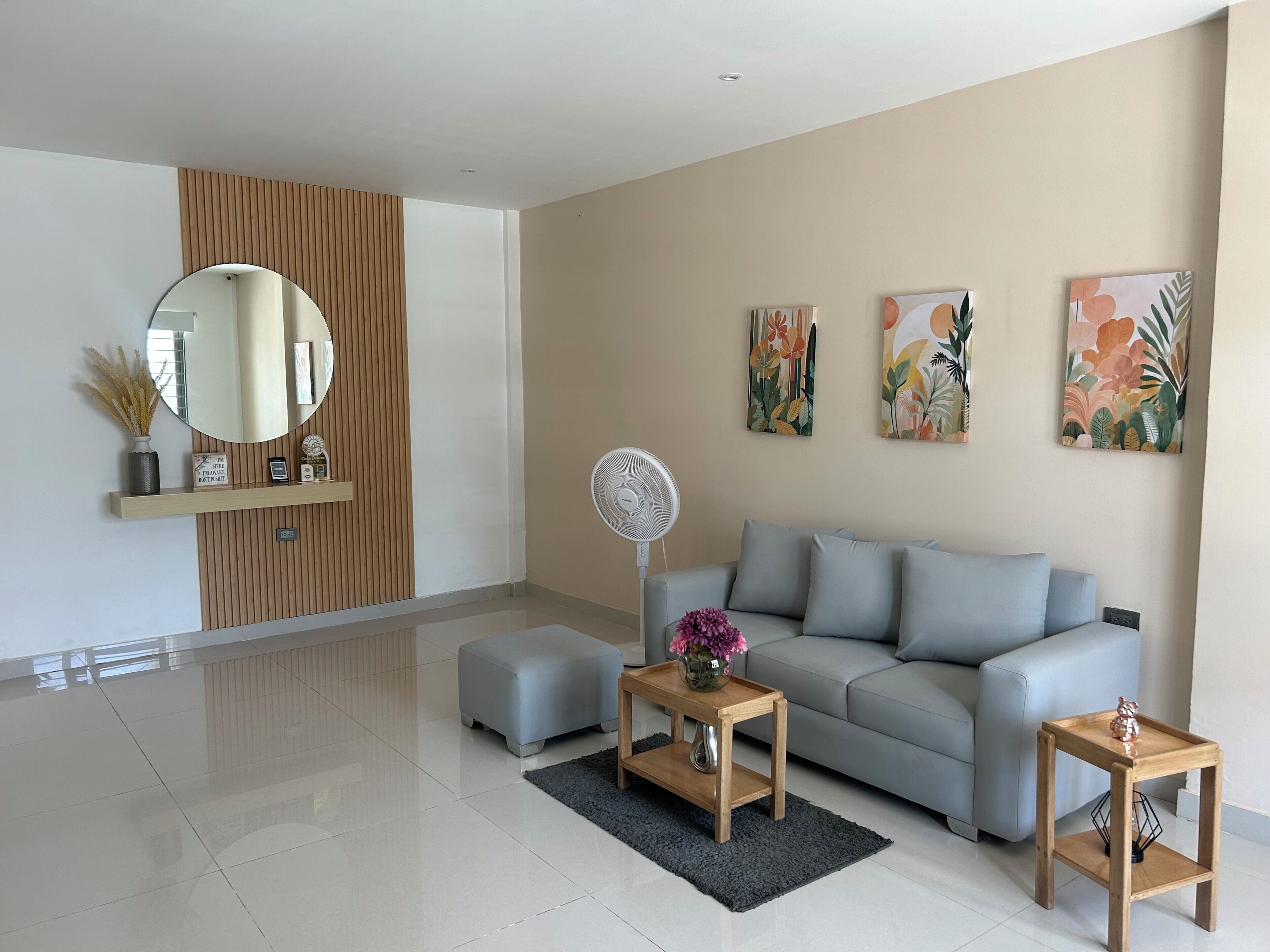 A modern living room with a grey sofa, a round mirror, a small wooden table with a vase, and a standing fan.