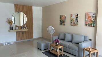 A modern living room with a grey sofa, a round mirror, a small wooden table with a vase, and a standing fan.