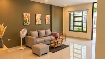 A modern living room with a sofa, ottoman, and a fan. There are paintings on the wall and a window with blinds.
