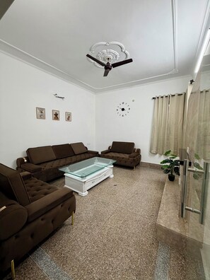 A living room with two brown sofas, a glass coffee table, a ceiling fan, a clock, and a potted plant.