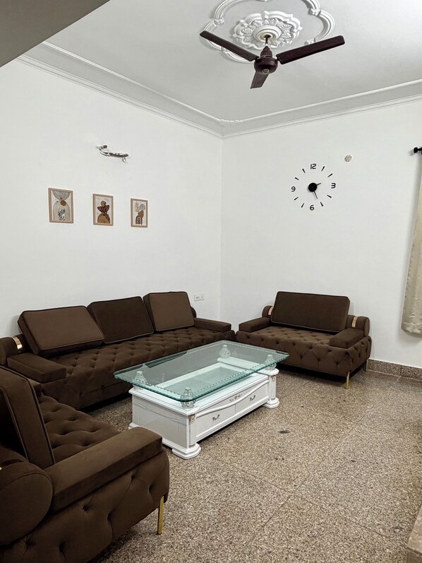 A living room with a brown sectional sofa, a glass coffee table, a ceiling fan, and a wall clock.