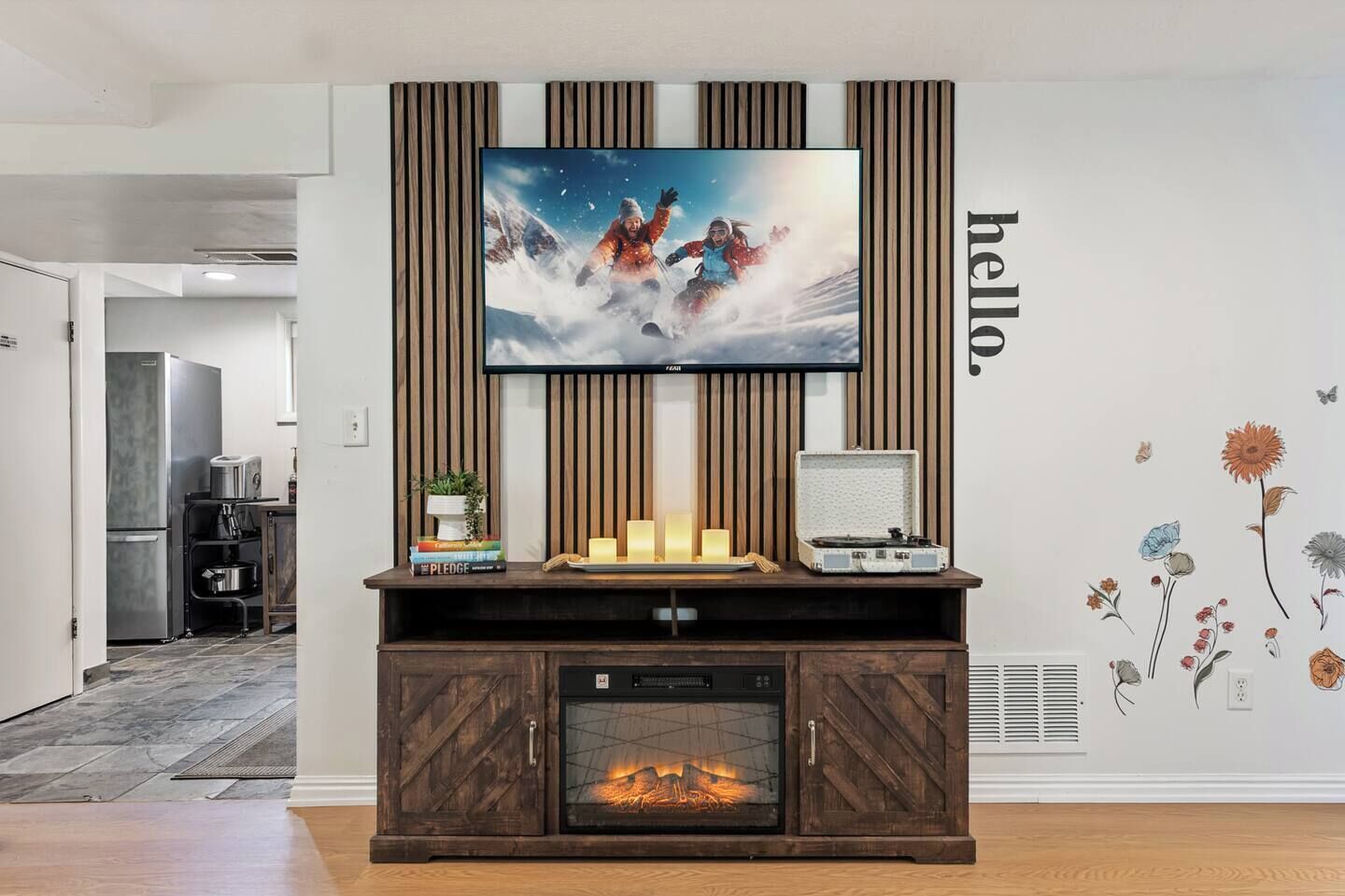 Een moderne woonkamer met een flatscreen televisie aan een houten paneel, een open haard onder, en een decoratieve wand met bloemstickers.