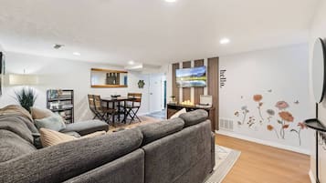 A modern living room with a sectional sofa, a dining area with a wooden table and chairs, a flat-screen TV mounted on the wall, and a decorative wall with floral decals.