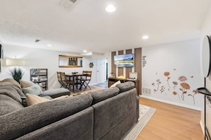 A modern living room with a sectional sofa, a dining area with a wooden table and chairs, a flat-screen TV mounted on the wall, and a decorative wall with floral decals.
