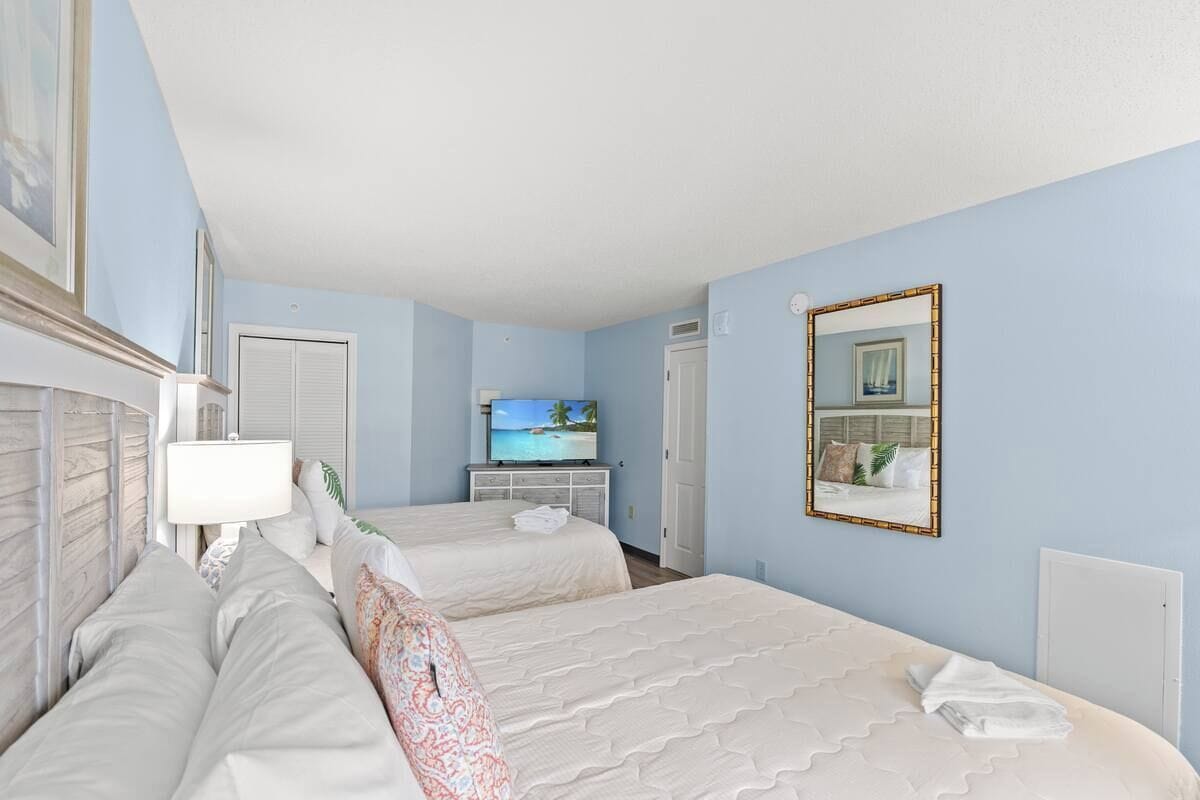 A hotel room with two beds, a television, a mirror, and a window with a view of the beach.