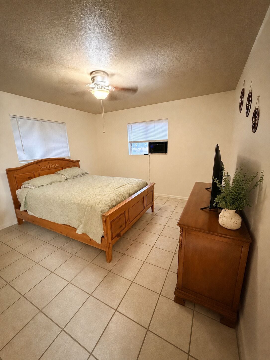 Une chambre à coucher avec un lit en bois, un ventilateur de plafond, une petite table avec une plante et un téléviseur fixé au mur.