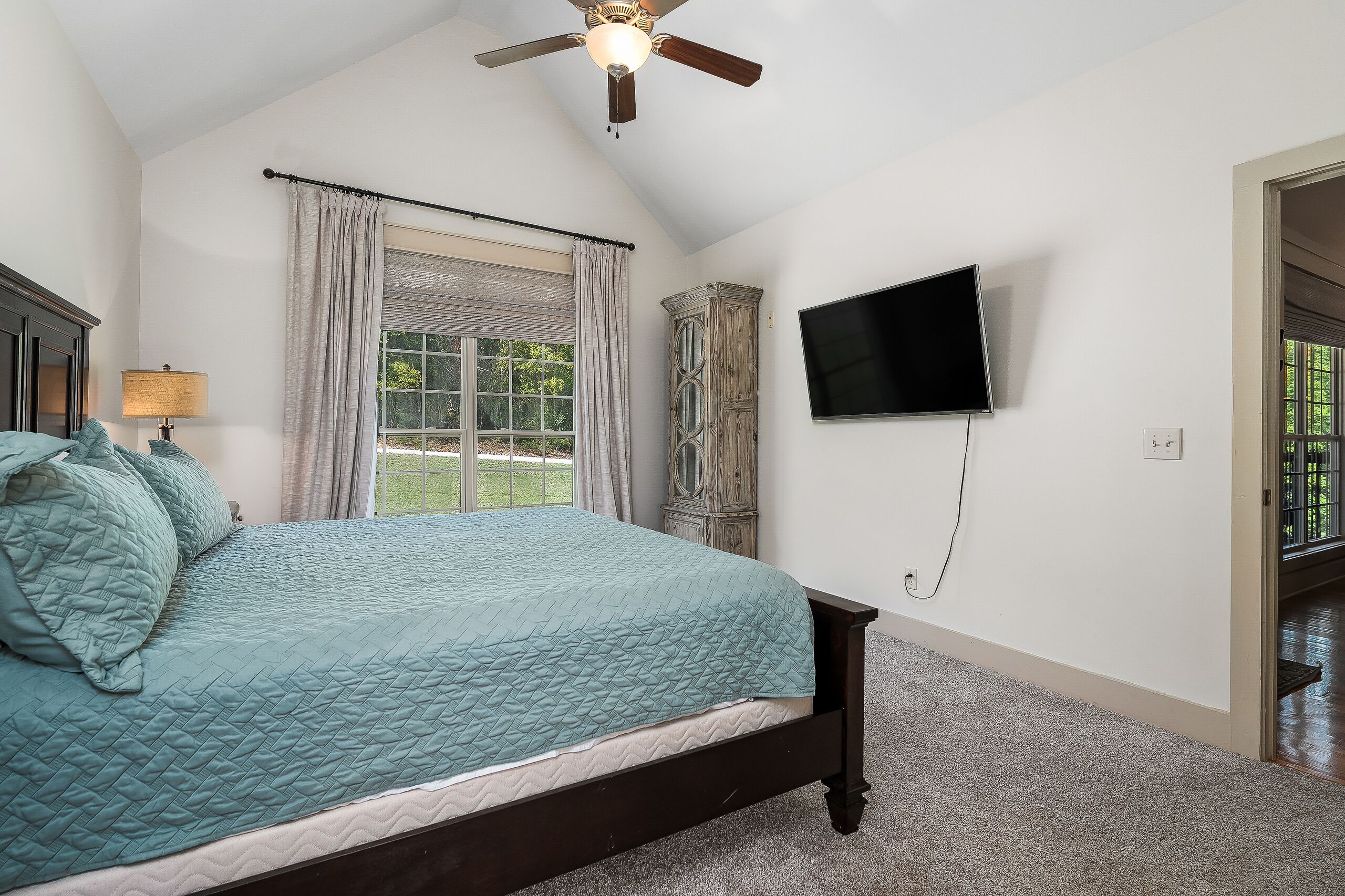 A bedroom with a bed, a ceiling fan, a window with curtains, a television mounted on the wall, and a door leading to another room.