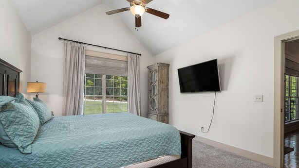 Un dormitorio con una cama, ventilador de techo, ventana con cortinas, televisión montada en la pared y una puerta que da a otra habitación.