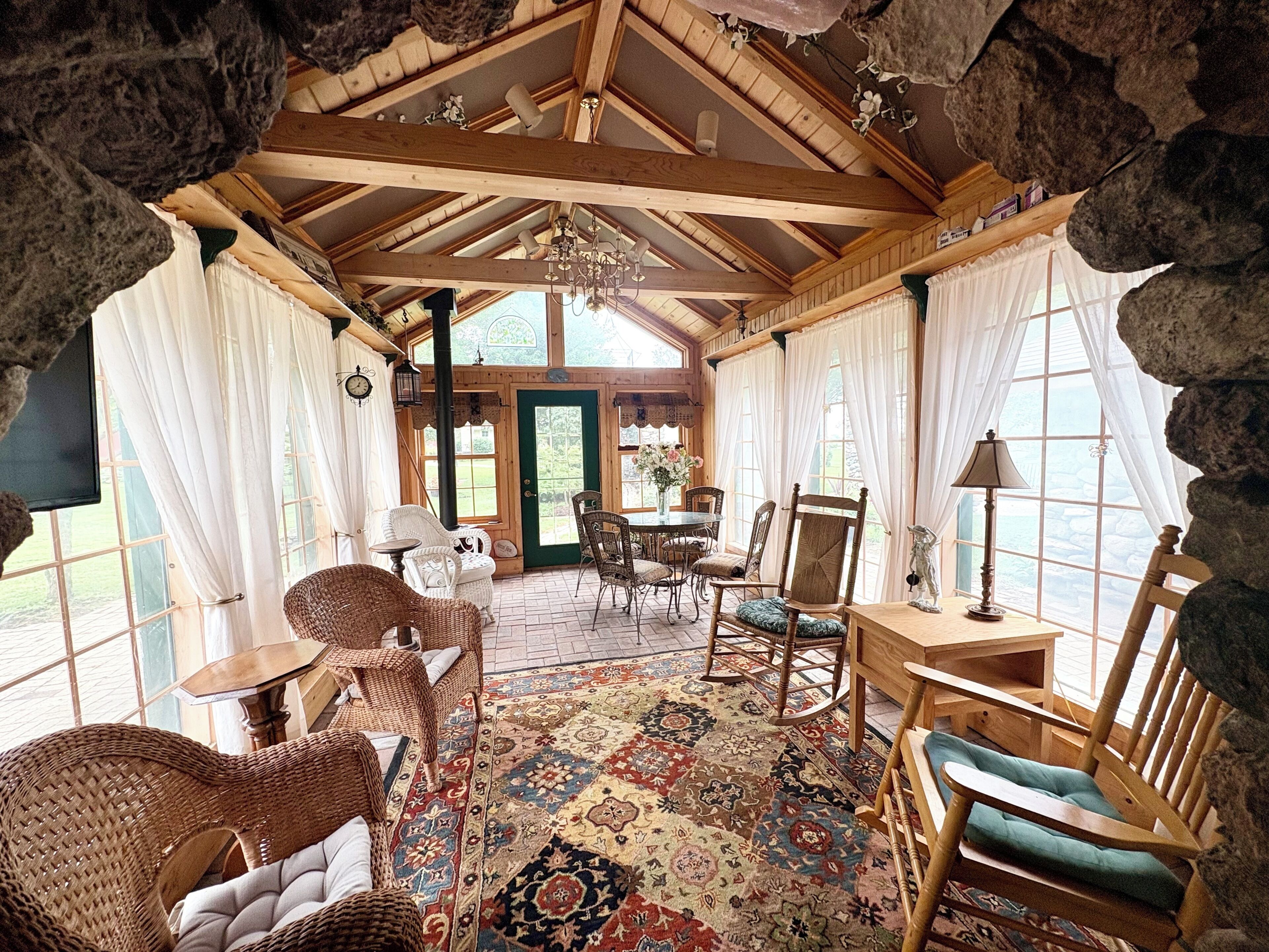 A rustic interior with a wooden ceiling, stone walls, a dining area with chairs and a table, and a patterned rug.