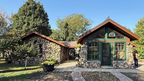 Un bâtiment en pierre avec une porte et des fenêtres vertes, entouré d’arbres et bordé d’un chemin pavé.