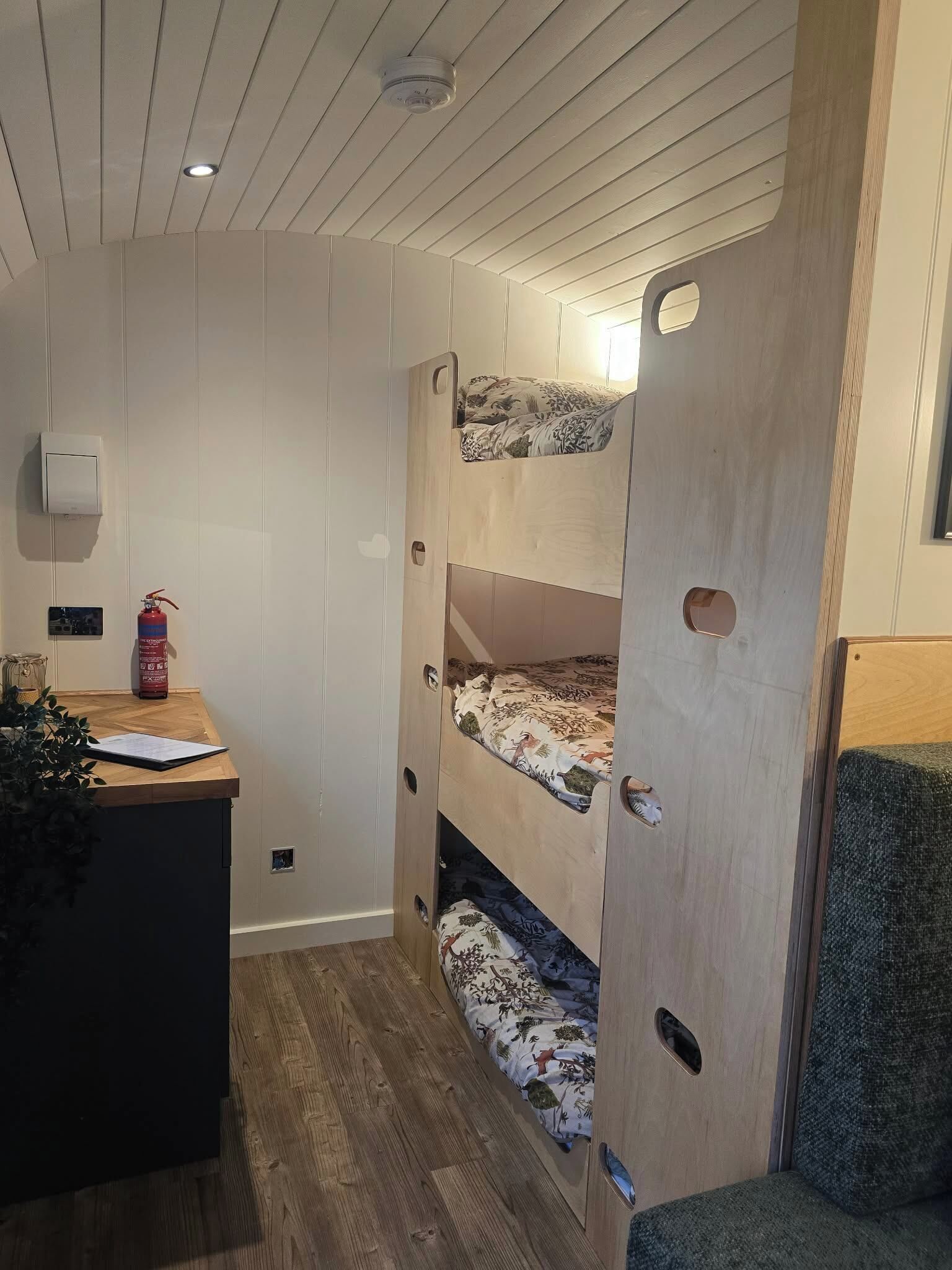 Een stapelbed met een houten hoofdeinde, een bureau met een plant, en een brandblusser.