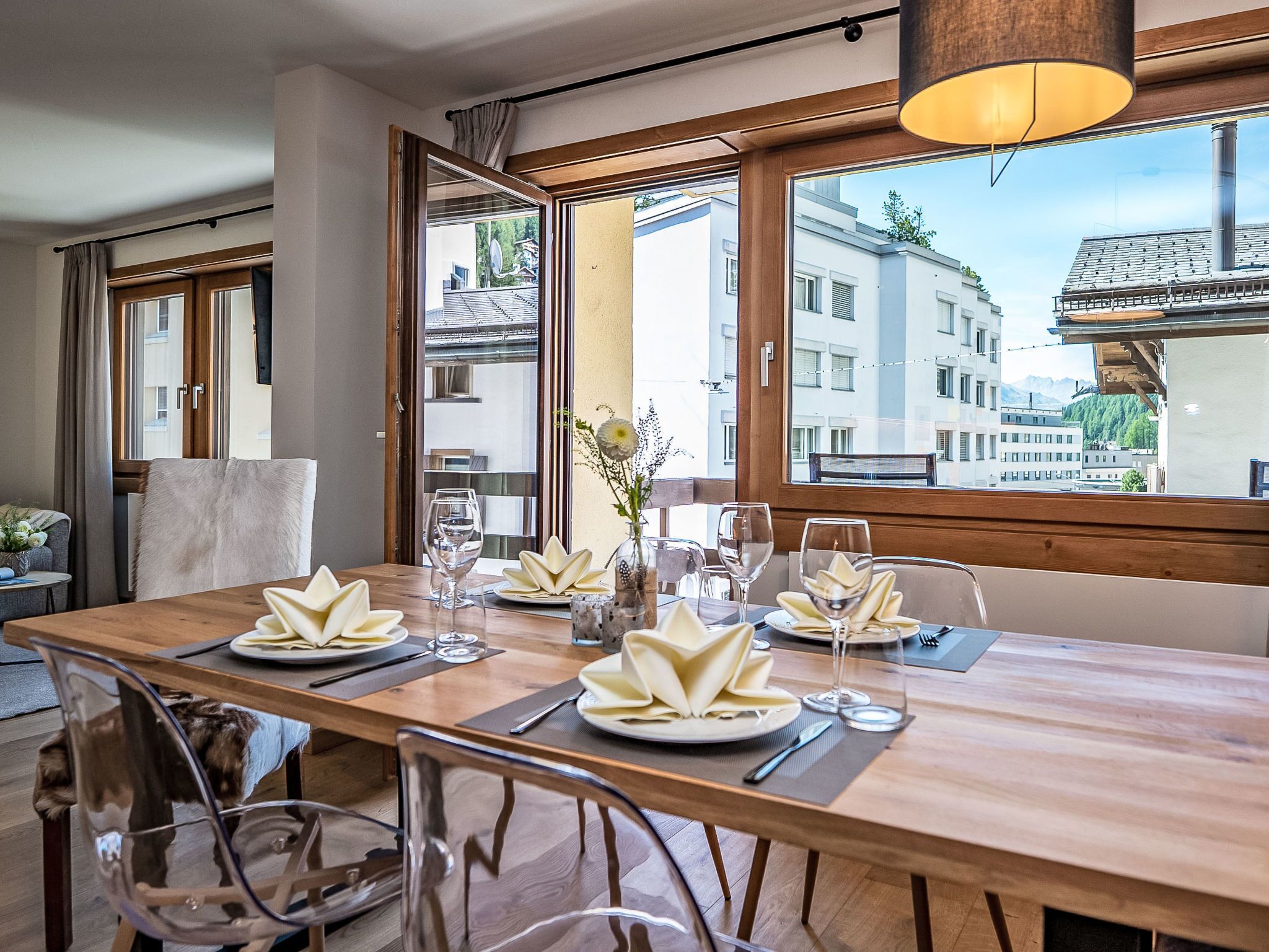 A dining area with a long wooden table set for four, large windows with a view of buildings, and a hanging light fixture.