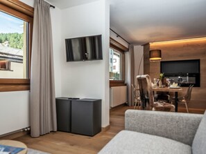 Ein modernes Wohnzimmer mit einem an der Wand befestigten Flachbildfernseher, einem Essbereich mit Tisch und Stühlen sowie einem großen Fenster mit Vorhängen.
