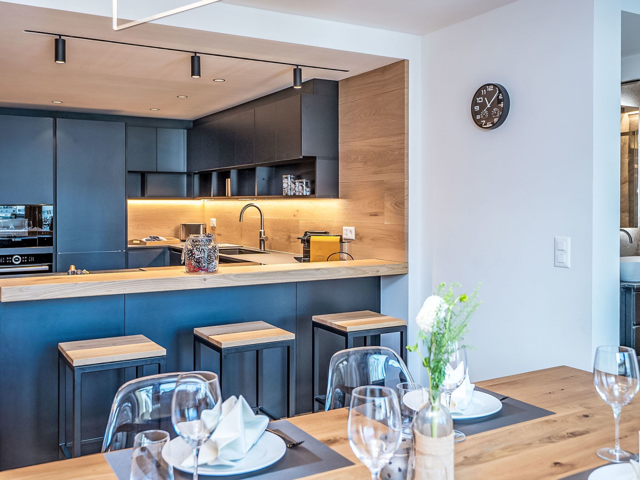 Une cuisine moderne avec des meubles de couleur sombre, un plan de travail en bois et un coin repas avec une table dressée pour deux personnes.