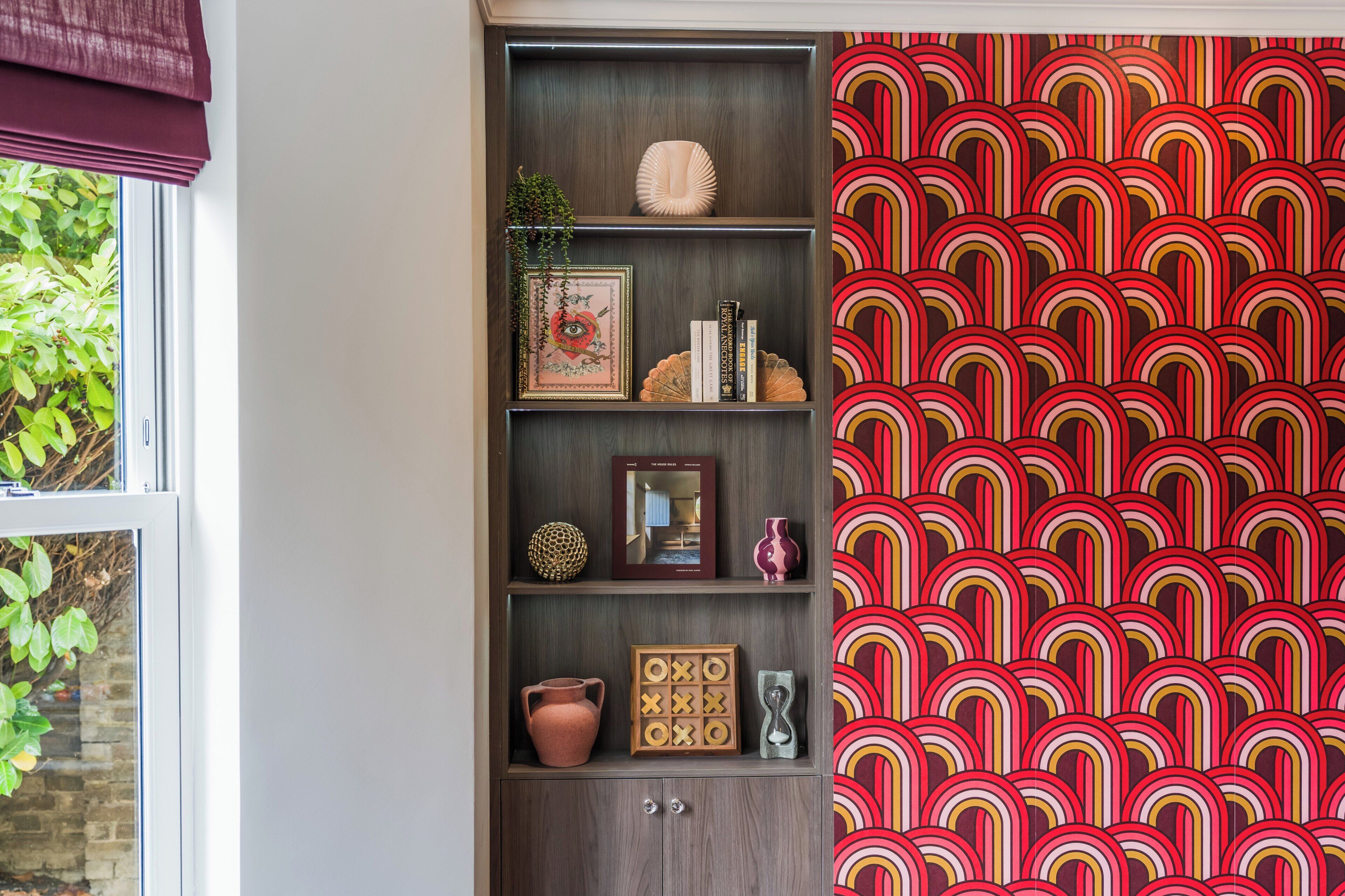 A room with a bookshelf containing various decorative items, a patterned wall, and a window with greenery outside.