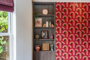 Een kamer met een boekenkast vol decoratieve objecten, een behang met patroon en een raam met beplanting erbuiten.