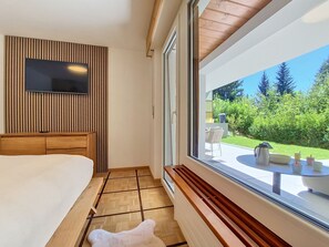 A bedroom with a bed, a TV mounted on a wooden panel, a window with a view of a patio, and a white pet on the floor. - Les Ambassadeurs B 3 by Interhome (Crans-Montana)