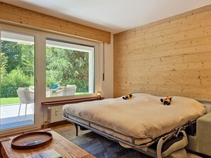 A bedroom with a large bed, a wooden headboard, a window with a view of greenery, and a small table with a bowl. - Les Ambassadeurs B 3 by Interhome (Crans-Montana)